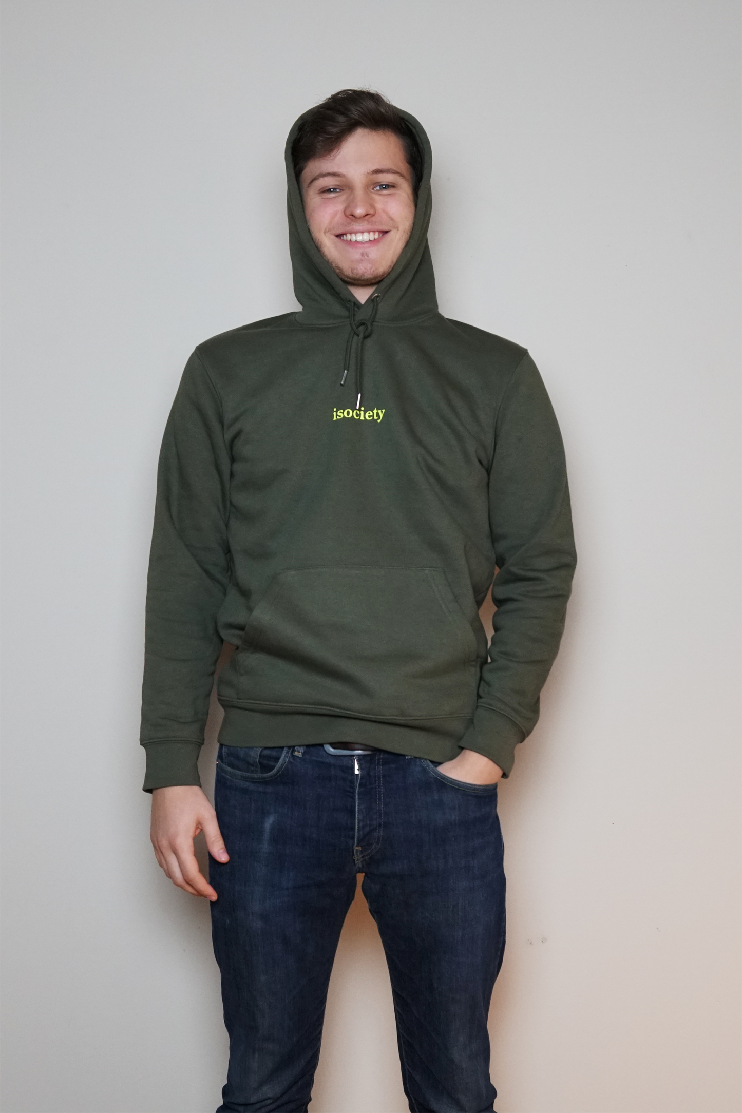 Ein Mann trägt dem Kapuzenpullover Noa und lacht in die Kamera. Er hat eine blaue Jeans an. Der Pulli ist Khaki. Auf der Brust steht in neon grün die Aufschrift isociety. Der Pulli hat vorne im unteren Bereich dieses für Kapuzenpulli typische Fach für die Hände. Hier auf dem Bild hat der Mann die Kapuze auf dem Kopf.