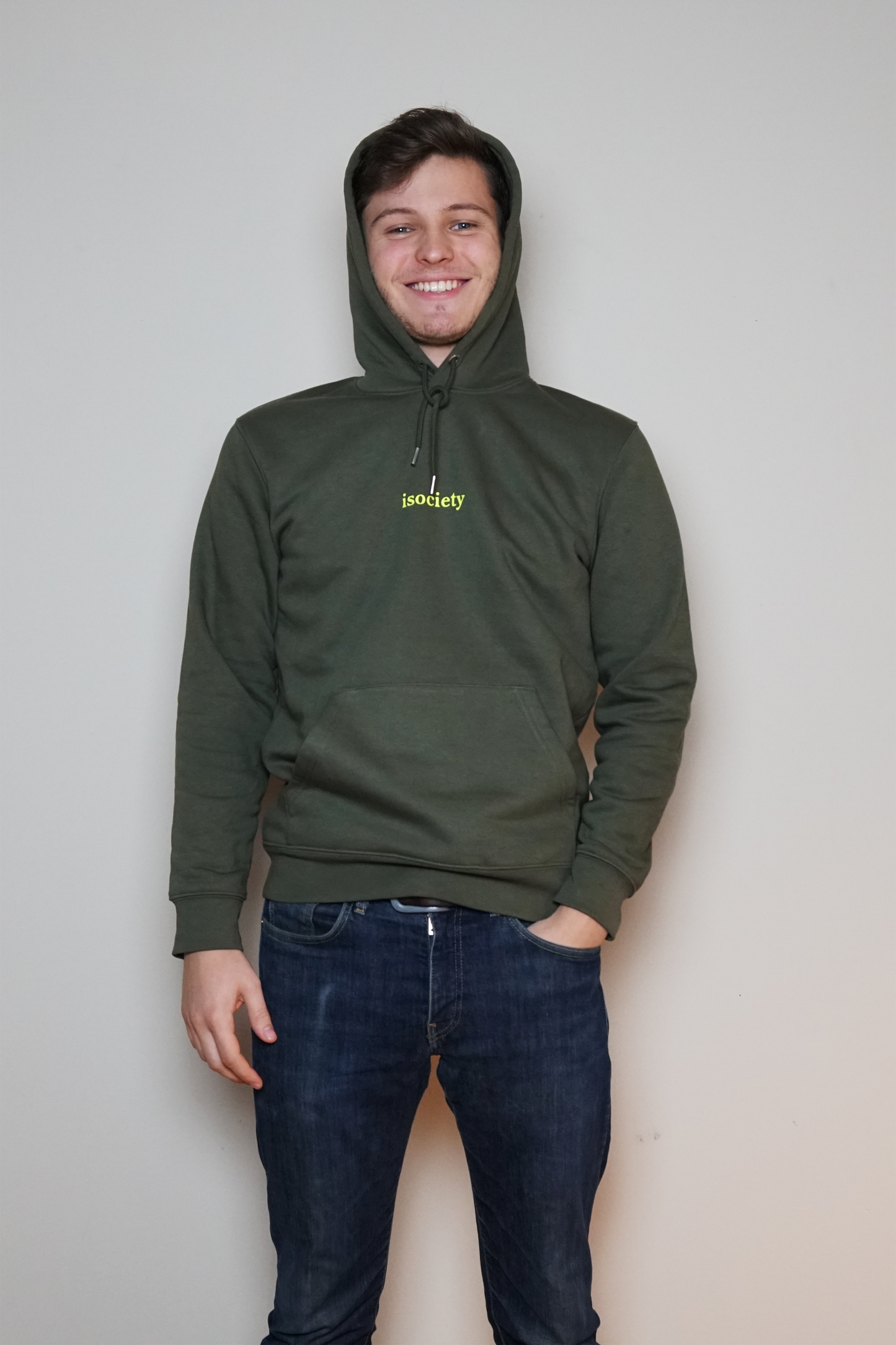 Ein Mann trägt dem Kapuzenpullover Noa und  lacht in die Kamera. Er hat eine blaue Jeans an. Der Pulli ist Khaki. Auf der Brust steht in neon grün die Aufschrift isociety. Der Pulli hat vorne im unteren Bereich dieses  für Kapuzenpulli typische Fach für die Hände. Hier auf dem Bild hat der Mann die Kapuze auf dem Kopf. 