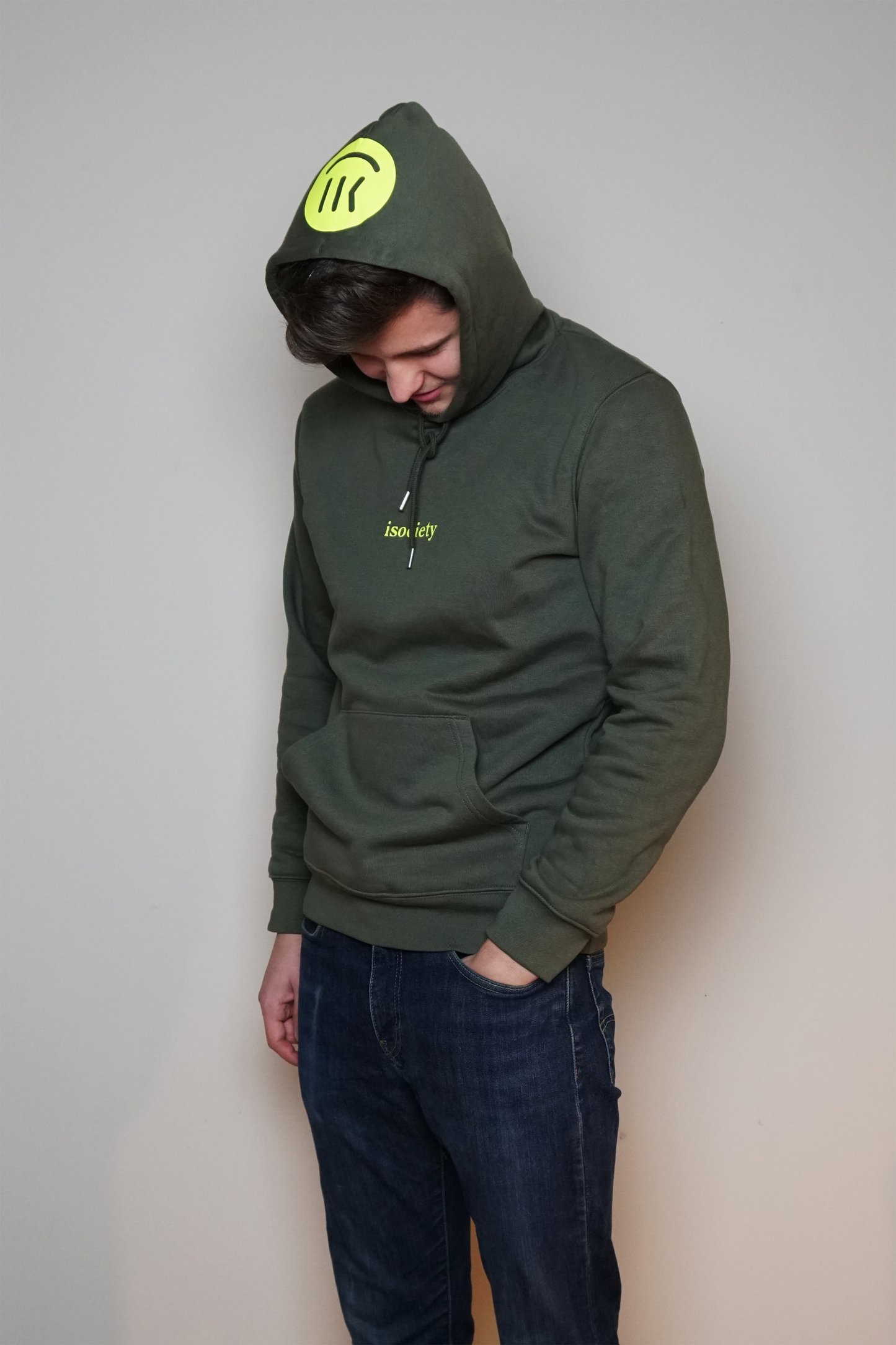 Ein Mann trägt den Kapuzenpulli mit dem Namen Noa in der Farbe Khaki. Er steht etwas seitlich und hat den Kopf nach unten geneigt. Der Kopf ist mit der Kapuze bedeckt. Auf der Kapuze ist der große isociety Smiley in neon grün zu erkennen.