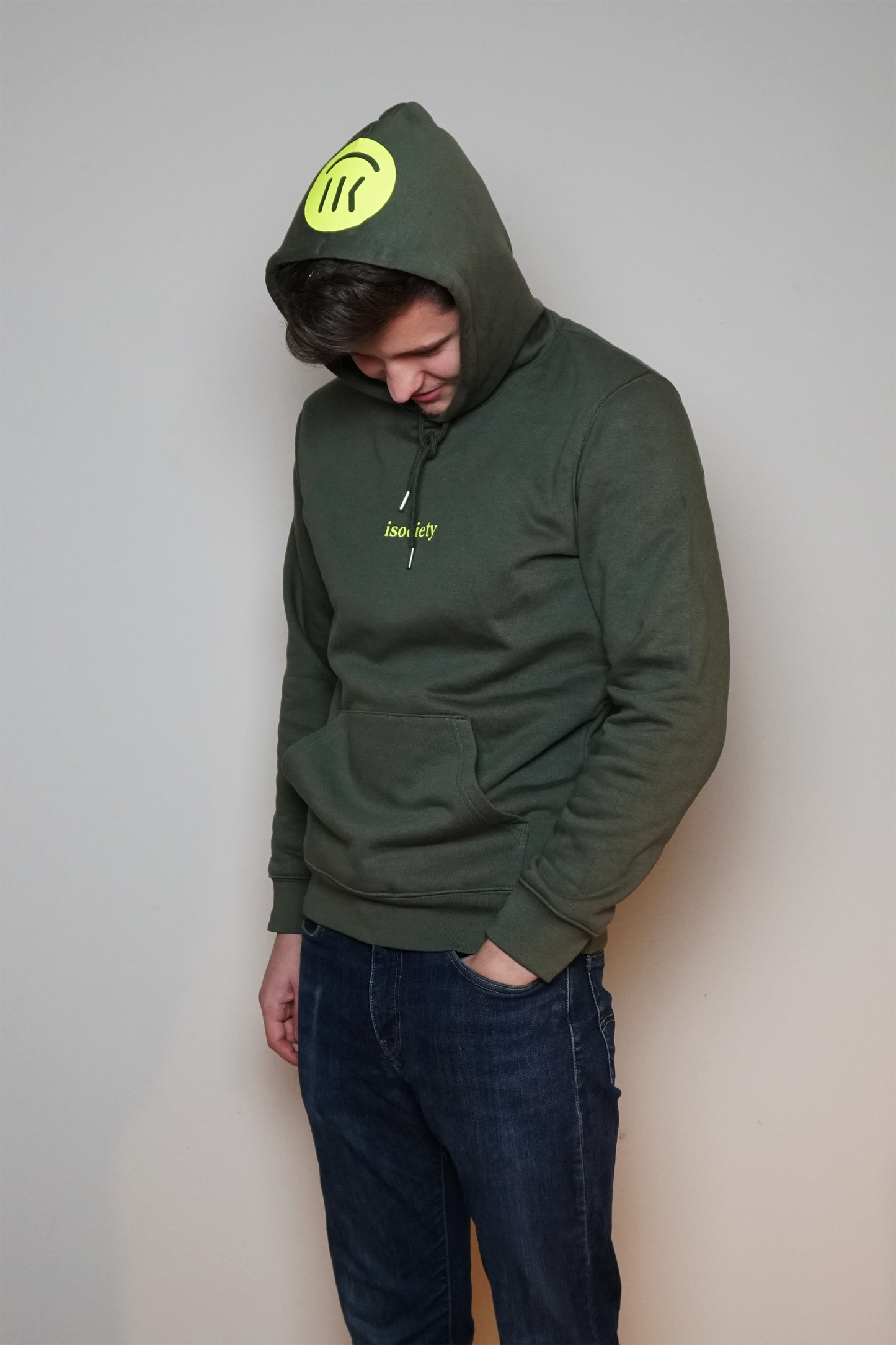 Ein Mann trägt den Kapuzenpulli mit dem Namen Noa in der Farbe Khaki. Er steht etwas seitlich und hat den Kopf nach unten geneigt. Der Kopf ist mit der Kapuze bedeckt. Auf der Kapuze ist der große isociety Smiley in neon grün zu erkennen. 