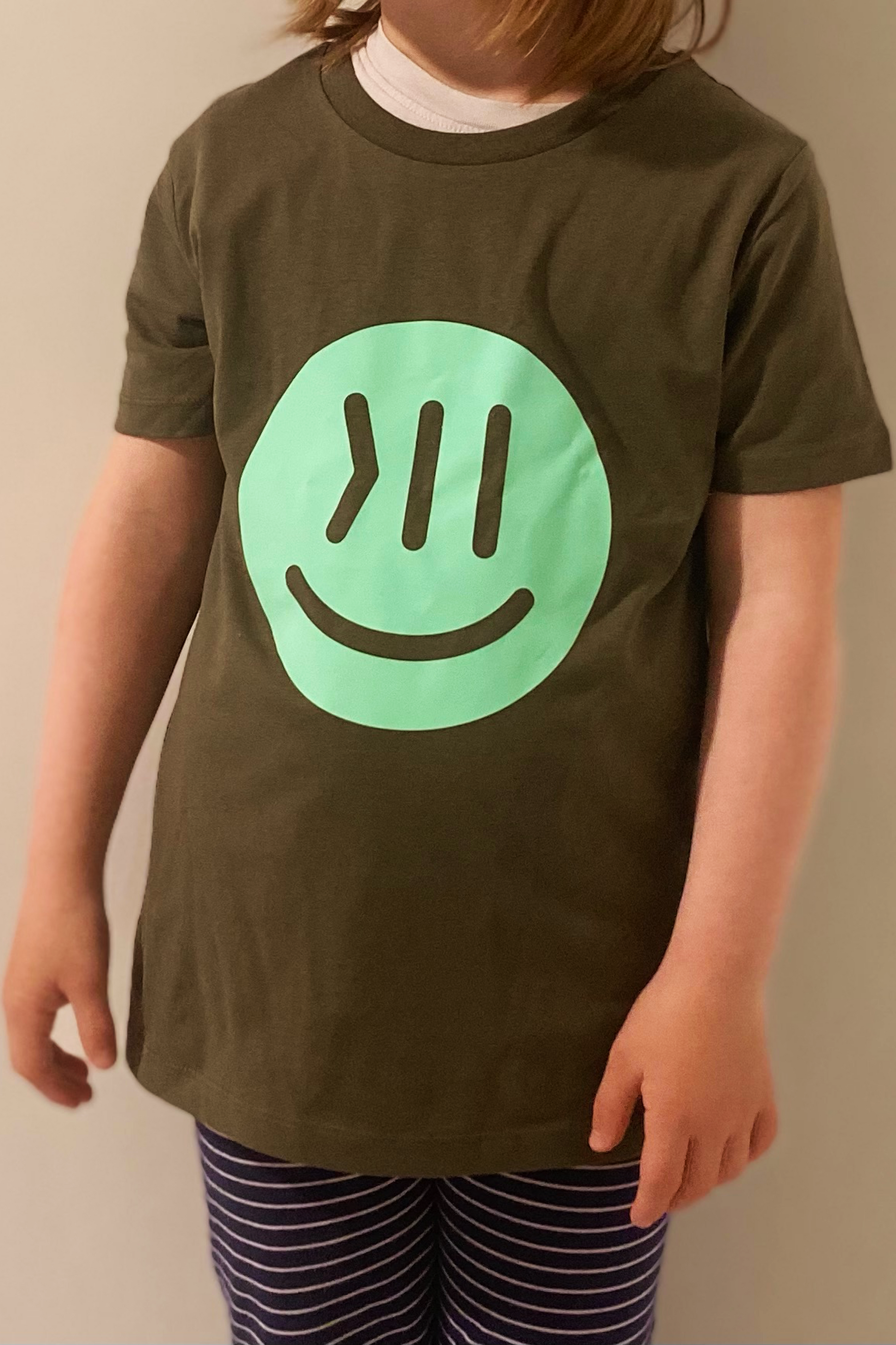 Ein Kind trägt das T-Shirt Juki in khaki mit dem isociety Smiley in mint. Das Kind ist vom Halsbereich bis zu den Knien zu sehen. Die Arme hängen lässig hinunter. Die Hose ist in blau mit dünnen weißen Streifen. Unter dem T-Shirt ist am Halsbereich zu erkennen, trägt das Kind ein weißes Unterhemd. Die Haare sind schulterlang und braun .
