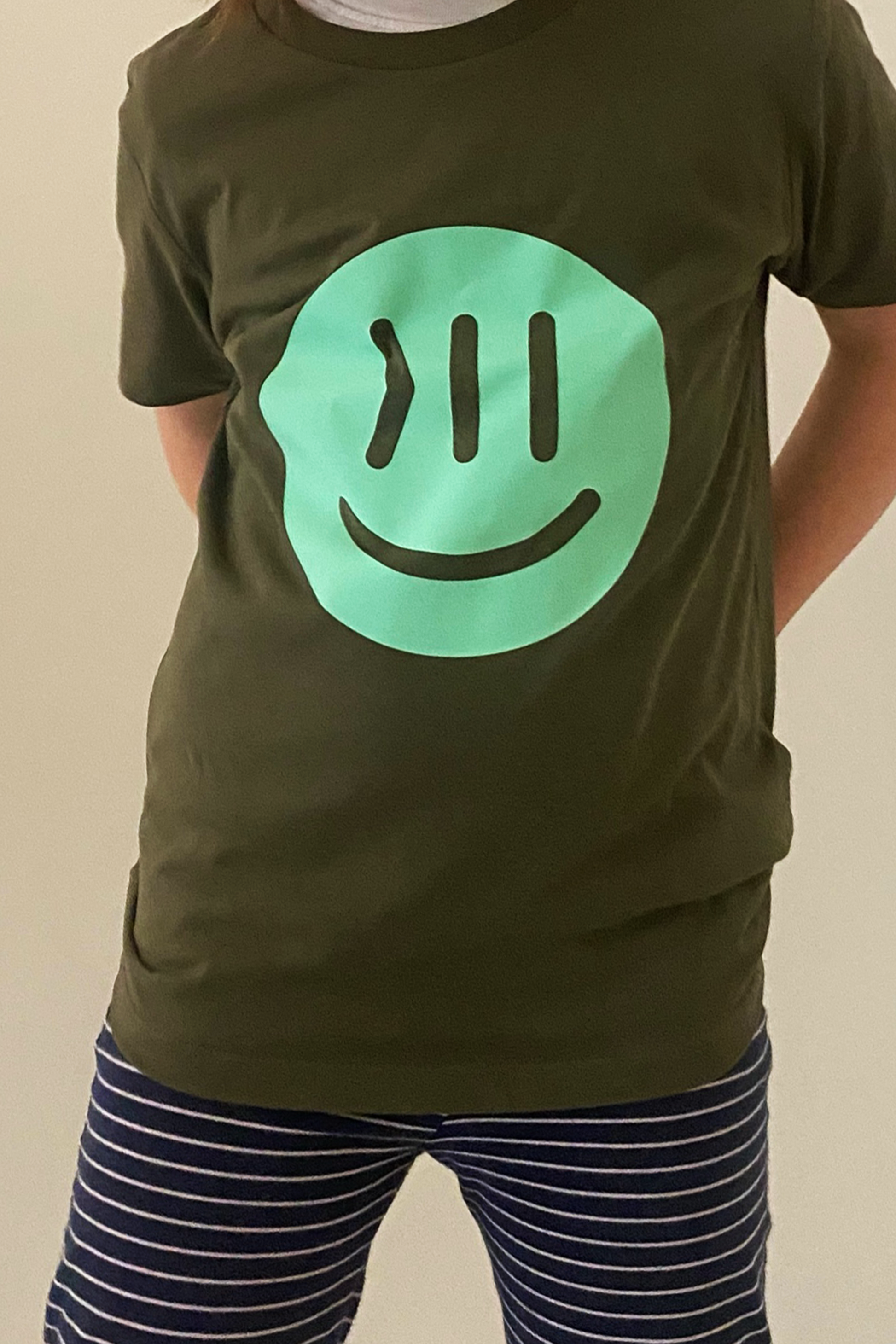 Ein Kind trägt das T-Shirt Juki in khaki mit dem isociety Smiley in mint. Das Kind ist vom Hals bis zu den Oberschenkeln zu sehen. Der Hintergrund ist weiß. Es sieht aus, als wenn das Kind  sein Gewicht auf das rechte Bein verlagert. Die Hände sind hinter dem Rücken, am sieht etwas die Ellenbogen. Die Hose ist in blau mit dünnen, weißen Streifen.