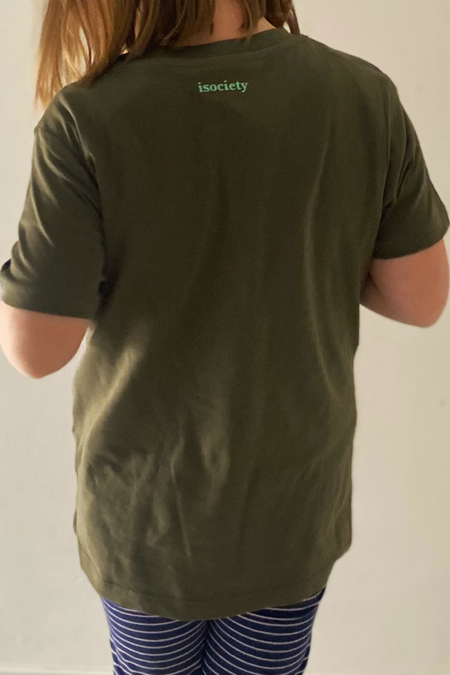 Das Kind trägt das T-Shirt Juki in khaki. Das Kind ist circa sieben Jahre alt und von hinten zu sehen. Man sieht den Bereich vom unteren Hinterkopf bis zu den Oberschenkeln. das Kind ist leicht nach links gedreht, die Beine schienen etwas überkreuz zu stehen. Im Nackenbereich ist die Aufschrift isociety in mint gedruckt. Das Kind hat mittellange braune Haare und eine lässige Hose in blau mit weißen Steifen. Der Hintergrund ist weiß.