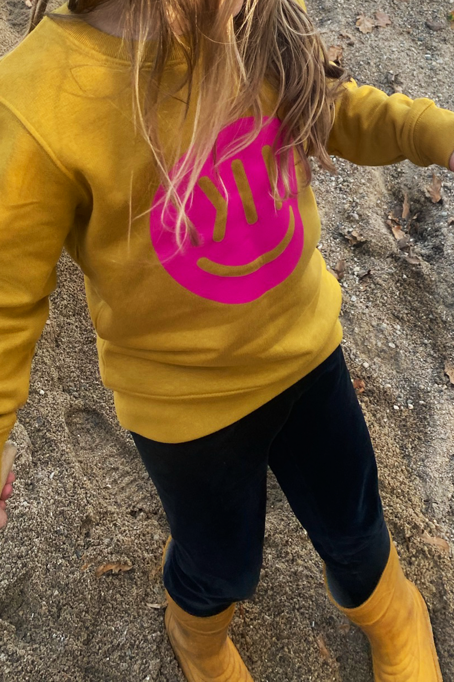 Ein Kind trägt den Sweater Kim. Das Kind ist von schräg oben fotografiert. Man sieht ungefähr den Bereich vom Hals bis zu den Füßen. Das Kind steht auf grobkörnigem Sand. Es trägt den Sweater Kim in ochre mit dem isociety Smiley in knalligem pink. Das Kind hat länge Haare, die offen hinunter hängen und zum Teil auf den Smiley fallen.Die Hose ist lässig und schwarz. Das Kind trägt Gummistiefel in gelb. Das Kind ist im Vorschulalter.