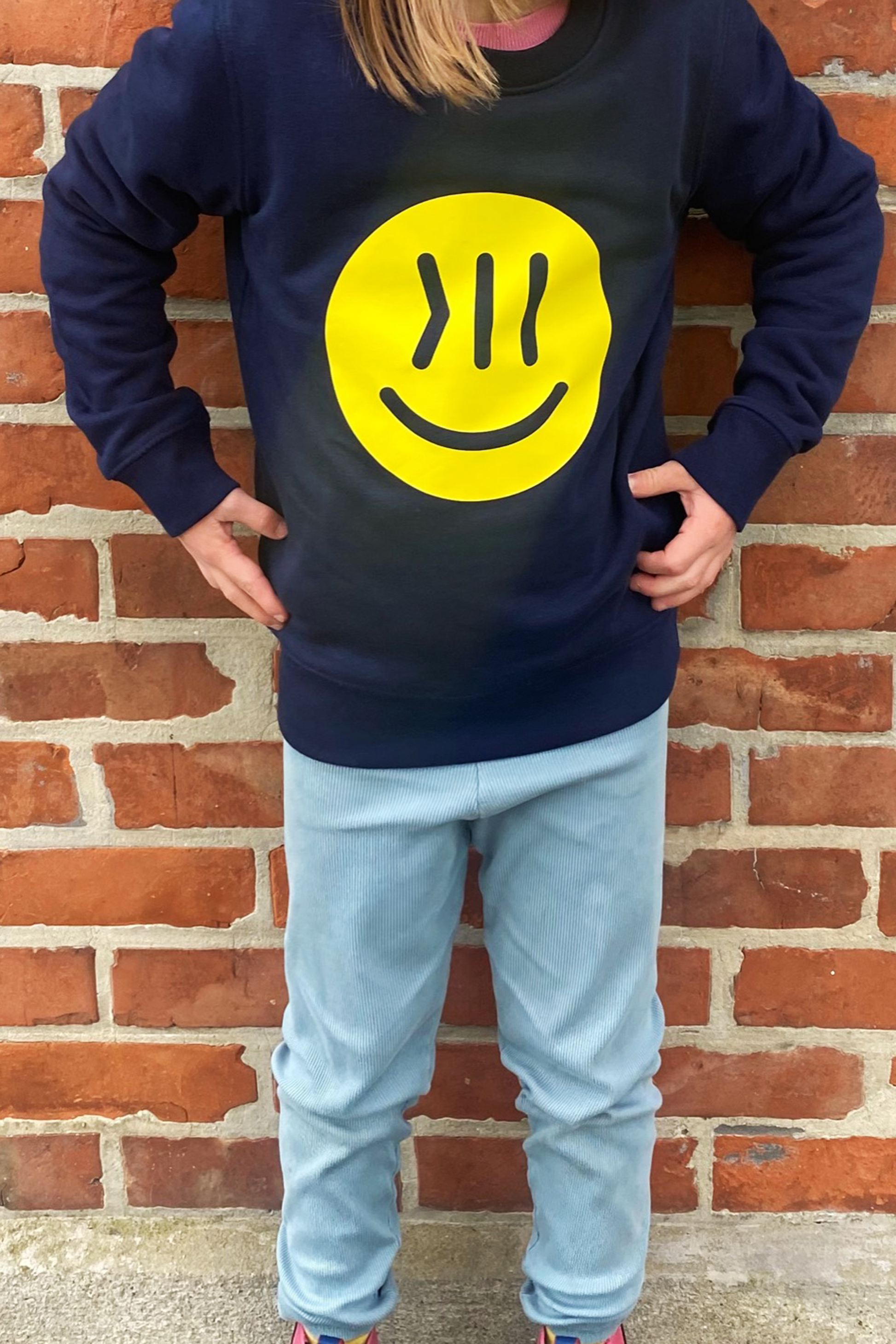 Ein Kind steht vor einer Wand. Der Kopf und die Füße des Kindes  sind nicht auf dem Bild zu sehen. Die Wand ist aus roten Backsteinen. Das Kind ist circa 7 Jahre. Es trägt den Pulli Elia. Das Kind ist von vorne zu sehen. Auf der Brust ist der große isociety Smiley in neon gelb zu sehen. Das Kind hält die Hände in die Hüften. Oben sieht man etwas die Haare ins Bild hängen, sie sind hellbraun. Das Kind trägt eine lässige Hose in hellblau.
