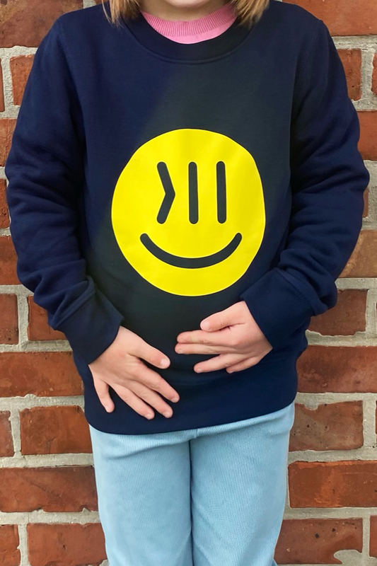 Ein Kind steht vor einer Wand. Der Kopf und die Füße des Kindes sind nicht auf dem Bild zu sehen.Man sieht das Kind bis zu den Knien. Die Wand ist aus roten Backsteinen. Das Kind ist circa 7 Jahre. Es trägt den Pulli Elia. Das Kind ist von vorne zu sehen. Auf der Brust ist der große isociety Smiley in neon gelb zu sehen. Das Kind legt die Hände vor den unteren Bauch. Oben sieht man etwas die Haare ins Bild hängen, sie sind hellbraun. Das Kind trägt eine lässige Hose in hellblau.